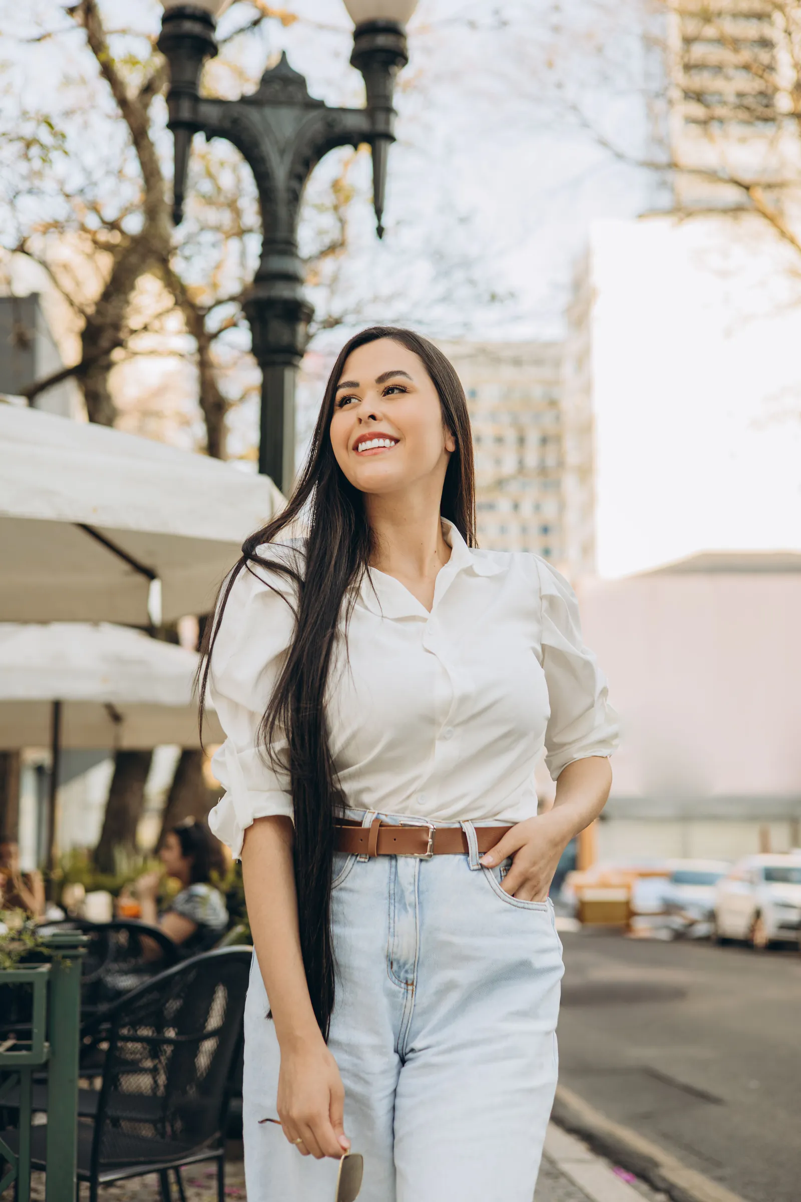 Femme brune souriante en chemise blanche et jean clair sur une terrasse, visuel d’ambiance